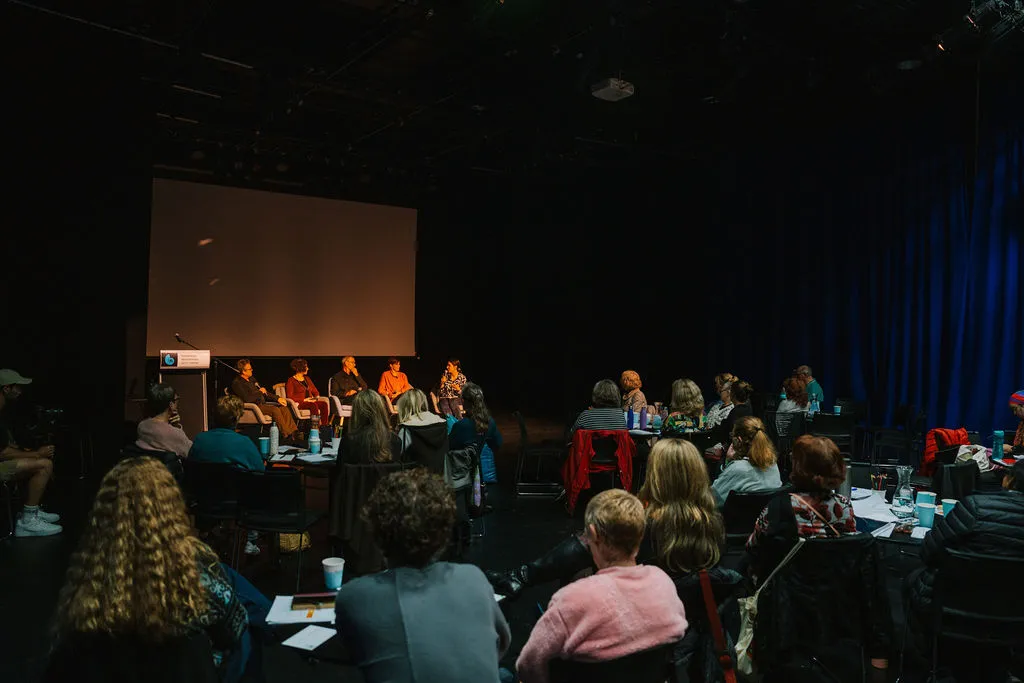 Audience seated in a dark theatre watching a panel of speakers on stage.